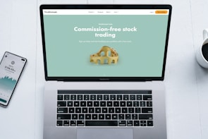 A laptop with a financial website open is centered on a white table. The screen displays a mint green background with text related to commission-free stock trading, accompanied by a gold object resembling a bridge. To the left, a smartphone displays a financial app with a graph and monetary figure. A cup of coffee sits on the right side, adding a casual work environment atmosphere.