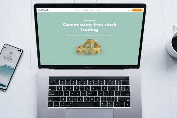 A laptop with a financial website open is centered on a white table. The screen displays a mint green background with text related to commission-free stock trading, accompanied by a gold object resembling a bridge. To the left, a smartphone displays a financial app with a graph and monetary figure. A cup of coffee sits on the right side, adding a casual work environment atmosphere.