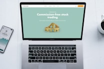 A laptop with a financial website open is centered on a white table. The screen displays a mint green background with text related to commission-free stock trading, accompanied by a gold object resembling a bridge. To the left, a smartphone displays a financial app with a graph and monetary figure. A cup of coffee sits on the right side, adding a casual work environment atmosphere.