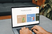 A person is typing on a MacBook Pro, viewing a website titled 'Personalize your portfolio.' The webpage displays three sections related to investing, each with different images and text. The laptop is on a grey table with a dark sofa and indoor plants in the background.