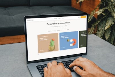 A person is typing on a MacBook Pro, viewing a website titled 'Personalize your portfolio.' The webpage displays three sections related to investing, each with different images and text. The laptop is on a grey table with a dark sofa and indoor plants in the background.