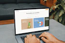 A person is typing on a MacBook Pro, viewing a website titled 'Personalize your portfolio.' The webpage displays three sections related to investing, each with different images and text. The laptop is on a grey table with a dark sofa and indoor plants in the background.