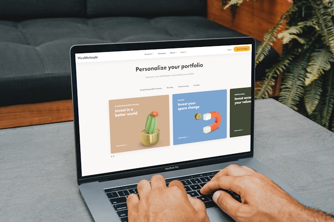 A person is typing on a MacBook Pro, viewing a website titled 'Personalize your portfolio.' The webpage displays three sections related to investing, each with different images and text. The laptop is on a grey table with a dark sofa and indoor plants in the background.