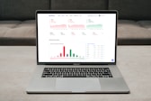 A laptop displaying a financial dashboard with various stock market graphs and data. The screen shows different stock indices and their performance, including line charts and a bar chart for market overview. The laptop is placed on a light-colored table, and a gray sofa is visible in the background.