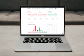 A laptop displaying a financial dashboard with various stock market graphs and data. The screen shows different stock indices and their performance, including line charts and a bar chart for market overview. The laptop is placed on a light-colored table, and a gray sofa is visible in the background.