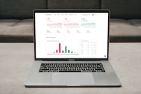 A laptop displaying a financial dashboard with various stock market graphs and data. The screen shows different stock indices and their performance, including line charts and a bar chart for market overview. The laptop is placed on a light-colored table, and a gray sofa is visible in the background.