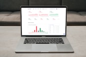 A laptop displaying a financial dashboard with various stock market graphs and data. The screen shows different stock indices and their performance, including line charts and a bar chart for market overview. The laptop is placed on a light-colored table, and a gray sofa is visible in the background.