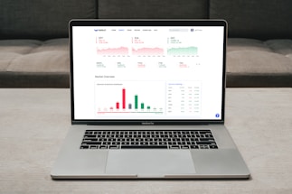 A laptop displaying a financial dashboard with various stock market graphs and data. The screen shows different stock indices and their performance, including line charts and a bar chart for market overview. The laptop is placed on a light-colored table, and a gray sofa is visible in the background.