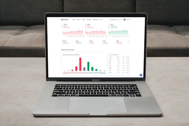 A laptop displaying a financial dashboard with various stock market graphs and data. The screen shows different stock indices and their performance, including line charts and a bar chart for market overview. The laptop is placed on a light-colored table, and a gray sofa is visible in the background.