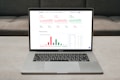 A laptop displaying a financial dashboard with various stock market graphs and data. The screen shows different stock indices and their performance, including line charts and a bar chart for market overview. The laptop is placed on a light-colored table, and a gray sofa is visible in the background.
