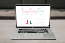 A laptop displaying a financial dashboard with various stock market graphs and data. The screen shows different stock indices and their performance, including line charts and a bar chart for market overview. The laptop is placed on a light-colored table, and a gray sofa is visible in the background.