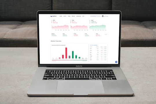 A laptop displaying a financial dashboard with various stock market graphs and data. The screen shows different stock indices and their performance, including line charts and a bar chart for market overview. The laptop is placed on a light-colored table, and a gray sofa is visible in the background.