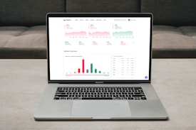 A laptop displaying a financial dashboard with various stock market graphs and data. The screen shows different stock indices and their performance, including line charts and a bar chart for market overview. The laptop is placed on a light-colored table, and a gray sofa is visible in the background.