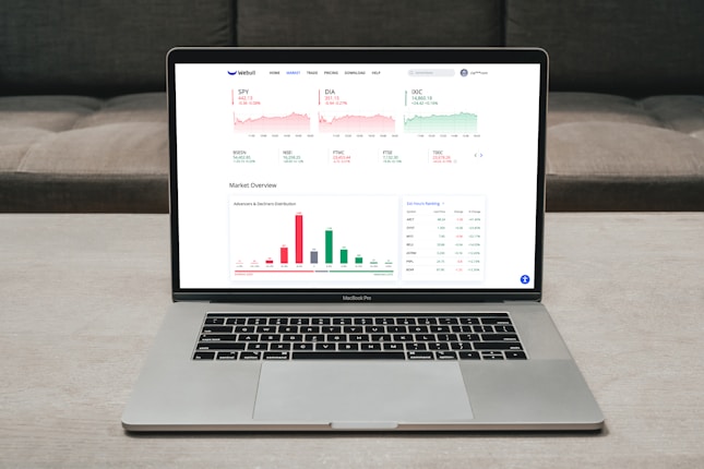 A laptop displaying a financial dashboard with various stock market graphs and data. The screen shows different stock indices and their performance, including line charts and a bar chart for market overview. The laptop is placed on a light-colored table, and a gray sofa is visible in the background.