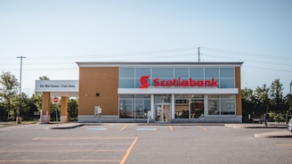 A standalone bank building with a modern design, featuring a light brick facade and a prominent red sign displaying the Scotiabank logo. There is a parking lot in front with marked parking spaces, and a sign that says 'Do Not Enter / Exit Only'. The area is surrounded by some trees and power lines are visible in the background.