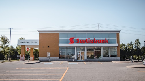 A standalone bank building with a modern design, featuring a light brick facade and a prominent red sign displaying the Scotiabank logo. There is a parking lot in front with marked parking spaces, and a sign that says 'Do Not Enter / Exit Only'. The area is surrounded by some trees and power lines are visible in the background.