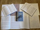 Several open books with printed text and musical sheets are spread out on a wooden surface. The central book is titled 'The Complete Oxford Shakespeare', and a pair of black-framed glasses rests on it. The surrounding pages contain a mixture of sheet music and text.