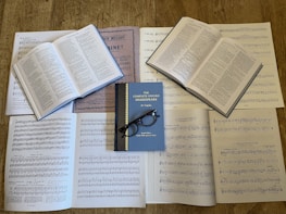 Several open books with printed text and musical sheets are spread out on a wooden surface. The central book is titled 'The Complete Oxford Shakespeare', and a pair of black-framed glasses rests on it. The surrounding pages contain a mixture of sheet music and text.