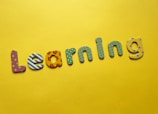 Colorful, decorative letters spelling 'Learning' are arranged across a bright yellow background. Each letter has a different pattern, including stripes, polka dots, flowers, and stars.