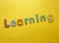 Colorful, decorative letters spelling 'Learning' are arranged across a bright yellow background. Each letter has a different pattern, including stripes, polka dots, flowers, and stars.