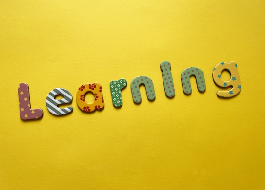 Colorful, decorative letters spelling 'Learning' are arranged across a bright yellow background. Each letter has a different pattern, including stripes, polka dots, flowers, and stars.