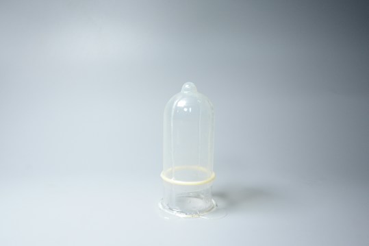 A transparent condom is placed upright against a plain light gray background. The image is minimalistic with the condom being the central focus.