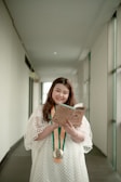 A person is standing in a hallway, wearing a white dress and holding a book while smiling. A medal with orange and green ribbons hangs around their neck. The hallway is bright, with white walls and large windows on one side.