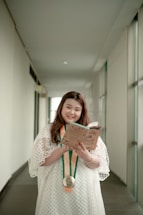 A person is standing in a hallway, wearing a white dress and holding a book while smiling. A medal with orange and green ribbons hangs around their neck. The hallway is bright, with white walls and large windows on one side.