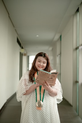 A person is standing in a hallway, wearing a white dress and holding a book while smiling. A medal with orange and green ribbons hangs around their neck. The hallway is bright, with white walls and large windows on one side.