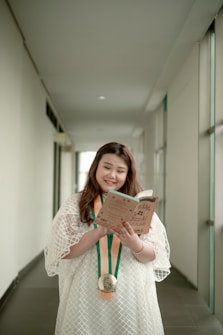 A person is standing in a hallway, wearing a white dress and holding a book while smiling. A medal with orange and green ribbons hangs around their neck. The hallway is bright, with white walls and large windows on one side.