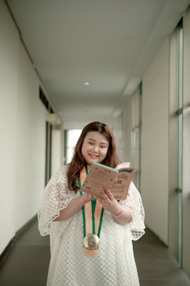 A person is standing in a hallway, wearing a white dress and holding a book while smiling. A medal with orange and green ribbons hangs around their neck. The hallway is bright, with white walls and large windows on one side.