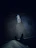 A small dog with glowing eyes stands on a concrete surface, illuminated in a dark environment. The perspective is from above, showing the feet and lower body of a person standing in front of the dog.