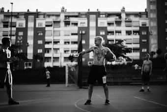 A group of young men are on an outdoor basketball court. They appear to be engaged in a casual game. The central figure with tattoos holds a basketball and looks towards two other shirtless men. An apartment building forms the backdrop, with several figures visible in the distance.