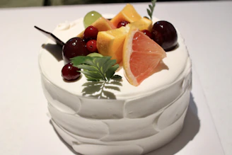 A pastel-colored ice cream cake being carefully decorated with fresh fruit and delicate frosting swirls.