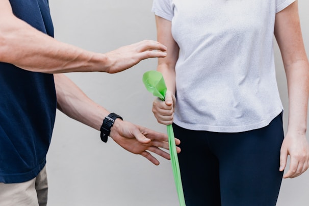 Two individuals are involved in an activity where one person is holding a green exercise band while the other appears to be guiding or instructing them. The person holding the band is wearing a white shirt and black pants, while the other person is in a dark shirt with a watch on their wrist.