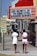A street scene featuring two women walking away from the camera. One wears a white dress, and the other sports a pink outfit. Above them is a bright red theater marquee with lights advertising 'Love Secrets of the Kama Sutra & Passion Seekers'. The sidewalk is bustling with pedestrians, and a 'No Right Turn' and 'One Way' sign is visible.