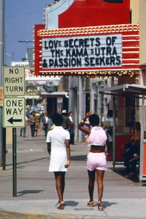 A street scene featuring two women walking away from the camera. One wears a white dress, and the other sports a pink outfit. Above them is a bright red theater marquee with lights advertising 'Love Secrets of the Kama Sutra & Passion Seekers'. The sidewalk is bustling with pedestrians, and a 'No Right Turn' and 'One Way' sign is visible.
