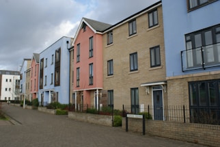 A row of modern, multi-story townhouses with different facade colors including blue, pink, and beige brick. The buildings feature large windows and small balconies, with a paved walkway and minimal landscaping in front. A cloudy sky forms the backdrop.