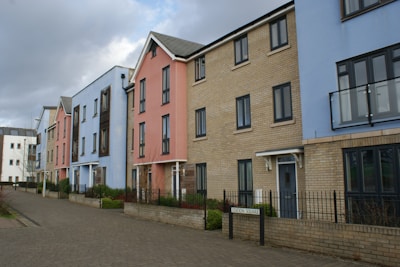 A row of modern, multi-story townhouses with different facade colors including blue, pink, and beige brick. The buildings feature large windows and small balconies, with a paved walkway and minimal landscaping in front. A cloudy sky forms the backdrop.