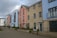 A row of modern, multi-story townhouses with different facade colors including blue, pink, and beige brick. The buildings feature large windows and small balconies, with a paved walkway and minimal landscaping in front. A cloudy sky forms the backdrop.