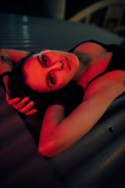 A person is lying down with arms crossed over their head, bathed in red light. The lighting creates dramatic shadows and highlights, giving a moody atmosphere. The background is out of focus, with a metallic surface beneath the person.