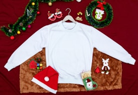 A white sweatshirt is laid out on a furry brown mat, surrounded by Christmas-themed decorations. These include a Santa hat, a pair of red novelty glasses with reindeer antlers, colorful ornaments, a 'Merry Christmas' sign, a small gift box, a green sock with Santa's face, a plush reindeer, and a wreath with a green bow. The background is a rich red fabric, adding to the festive atmosphere.