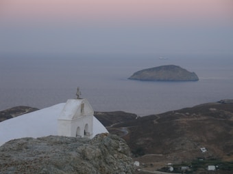 A tranquil seaside landscape featuring a small white chapel with a cross on its roof, perched on rocky terrain. In the background, a calm sea stretches out to meet a softly colored sky at sunset, with a distant island visible on the horizon.