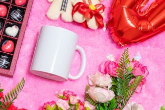 A white mug lies on a pink fluffy surface, surrounded by various decorative items. There is a heart-shaped red balloon and a plush teddy bear bow nearby. A box of chocolates with heart-shaped and round candies is placed to the left, and vibrant flowers with red, pink, and white blossoms are arranged at the bottom.