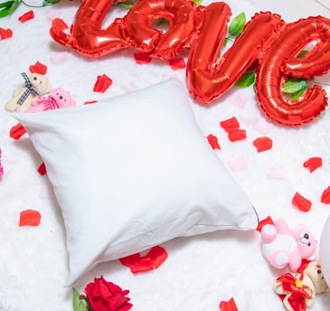 A white pillow is placed on a fluffy, white surface surrounded by scattered red and pink rose petals. Bright red, balloon-style letters spell out the word 'love' above the pillow. Small stuffed teddy bears in shades of pink and beige are positioned around the area, adding to the romantic and playful atmosphere.