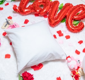 A white pillow is placed on a fluffy, white surface surrounded by scattered red and pink rose petals. Bright red, balloon-style letters spell out the word 'love' above the pillow. Small stuffed teddy bears in shades of pink and beige are positioned around the area, adding to the romantic and playful atmosphere.