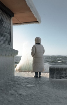 A person wearing a long, white winter coat with a hood stands on an icy surface, facing the sea. Strong winds create waves that splash dramatically against a nearby structure. The structure and ground are covered in thick layers of ice, suggesting extreme cold conditions.