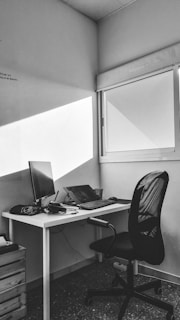 A black and white image of a minimalist office space featuring a desk with a computer monitor, keyboard, mouse, and some cables. A comfortable office chair is positioned nearby. Sunlight filters through a window, casting geometric light patterns on the wall.