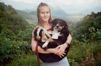 A person is holding a black and white dog in their arms while standing in a lush green landscape. The background features hills and dense vegetation, suggesting a natural rural setting. The person is smiling gently, and the dog appears calm and comfortable.