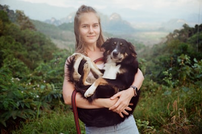 A person is holding a black and white dog in their arms while standing in a lush green landscape. The background features hills and dense vegetation, suggesting a natural rural setting. The person is smiling gently, and the dog appears calm and comfortable.